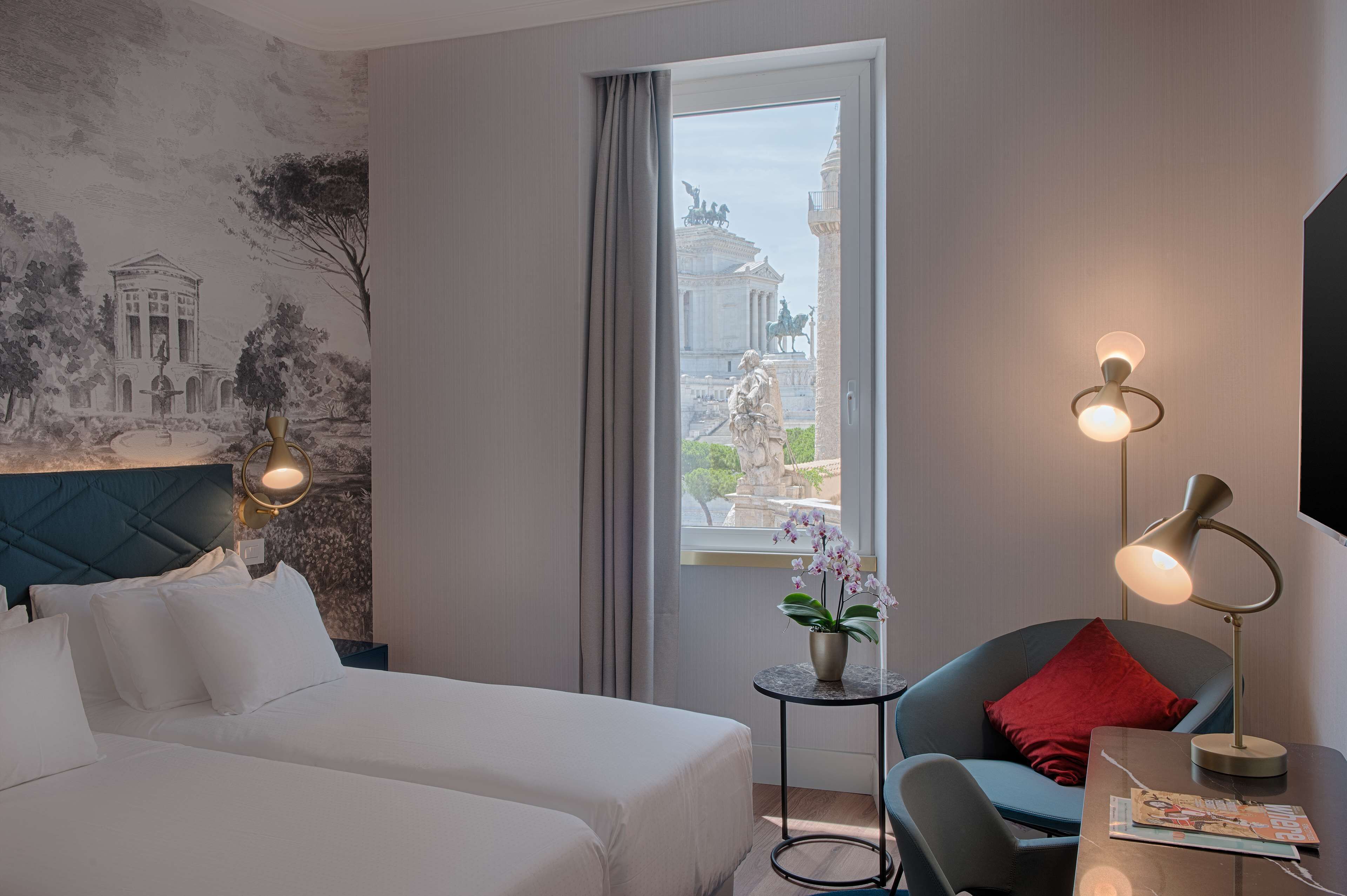 Una habitación de hotel con cama, una silla, una mesa, una lámpara, una ventana con vista a una estatua y un mural en la pared.