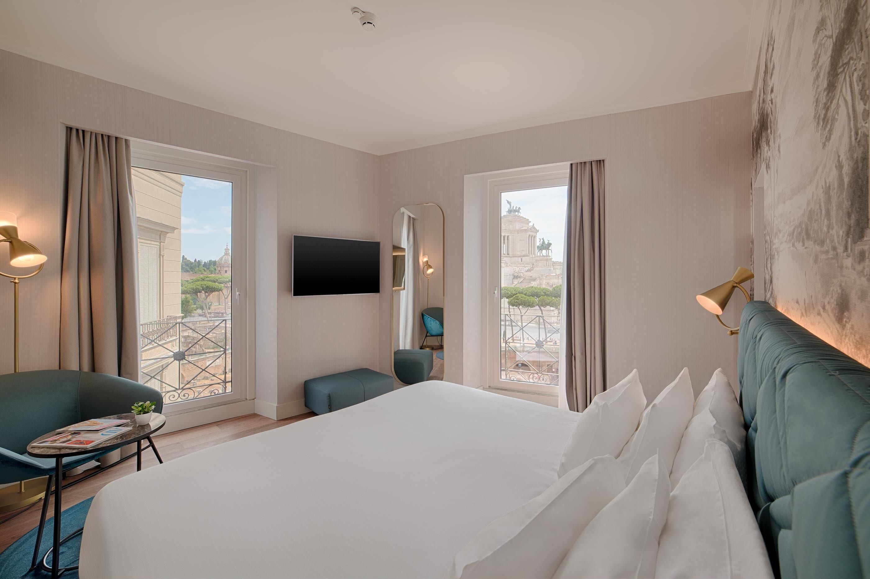 Ett hotellrum med en stor säng, en tv, en stol, ett litet bord med en växt och utsikt över en byggnad genom fönstret.