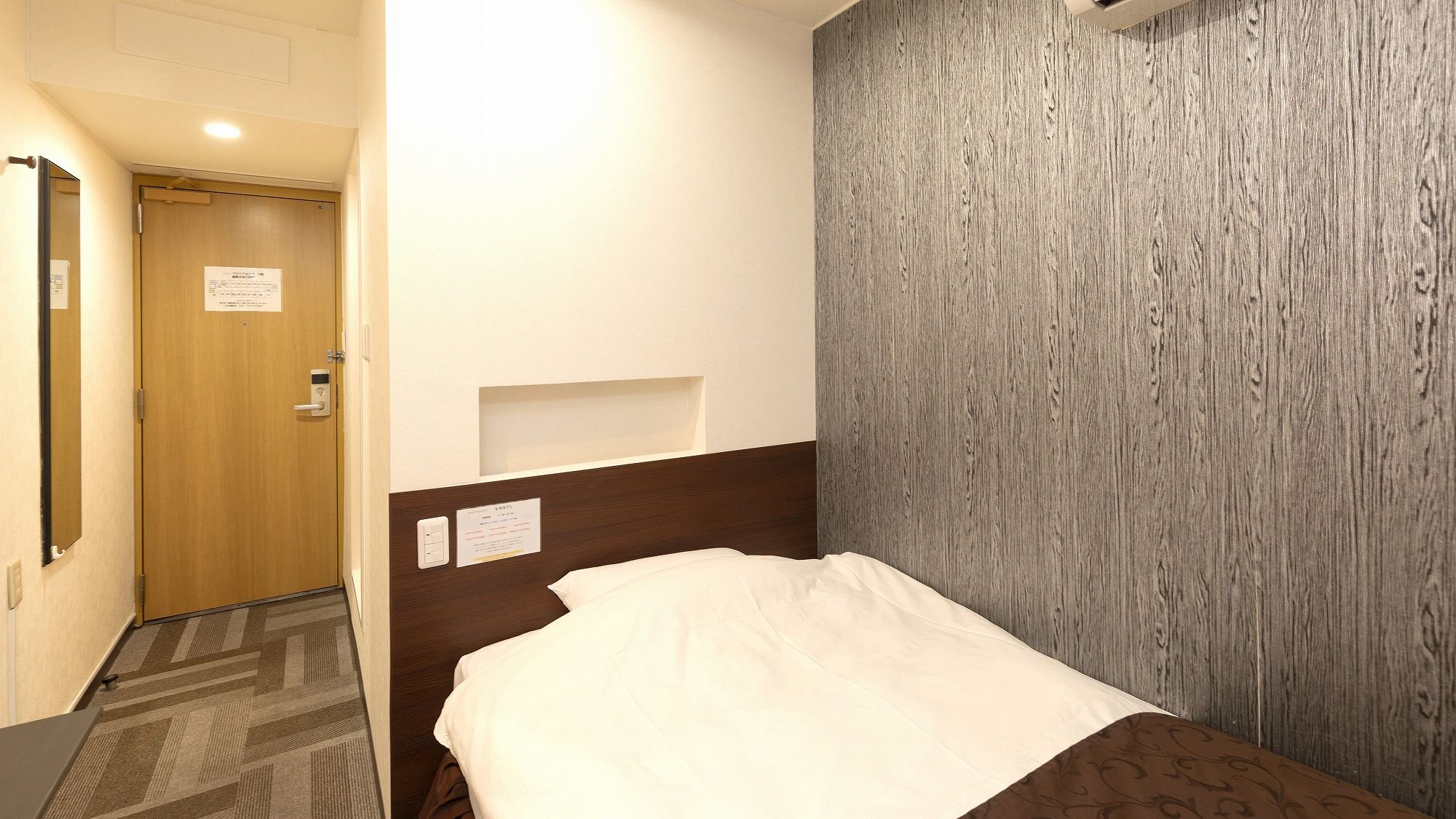 Une chambre d’hôtel avec une porte en bois, un lit avec un oreiller blanc, une étagère fixée au mur et un mur à texture.