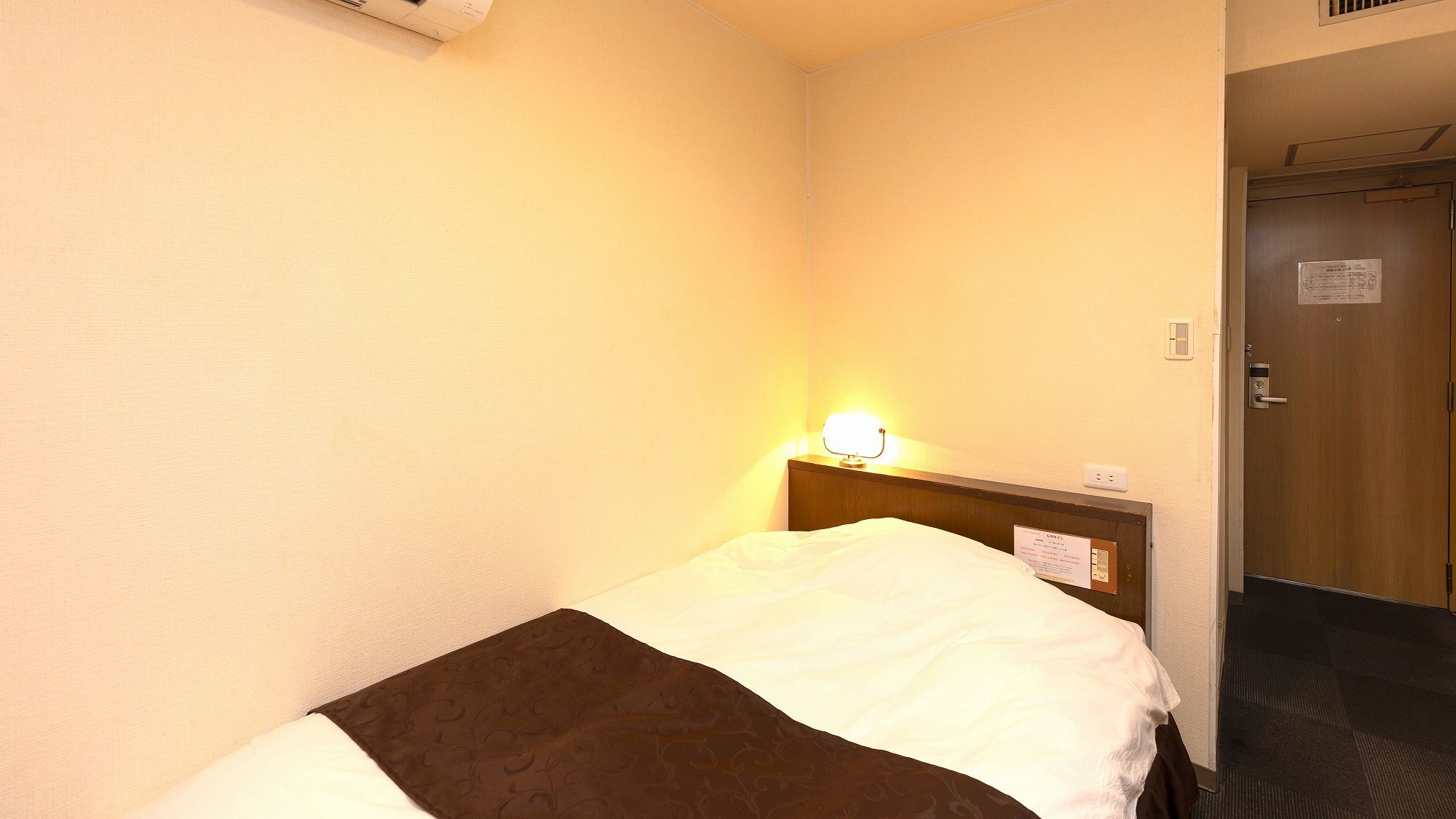Quarto de hotel com uma cama de solteiro, mesa de cabeceira, luminária na parede e porta com uma placa.