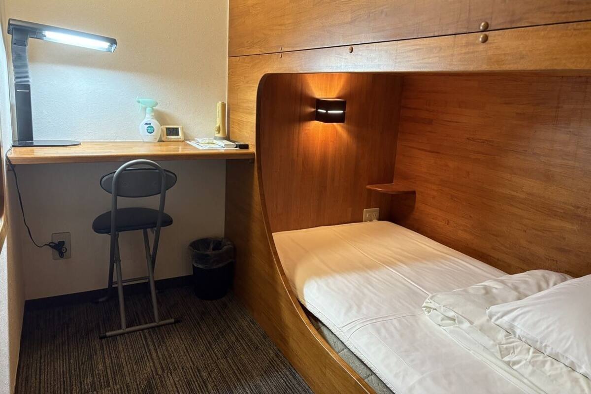 A compact room with a wooden headboard, a single bed, a desk with a chair, a desk lamp, and a small trash bin.