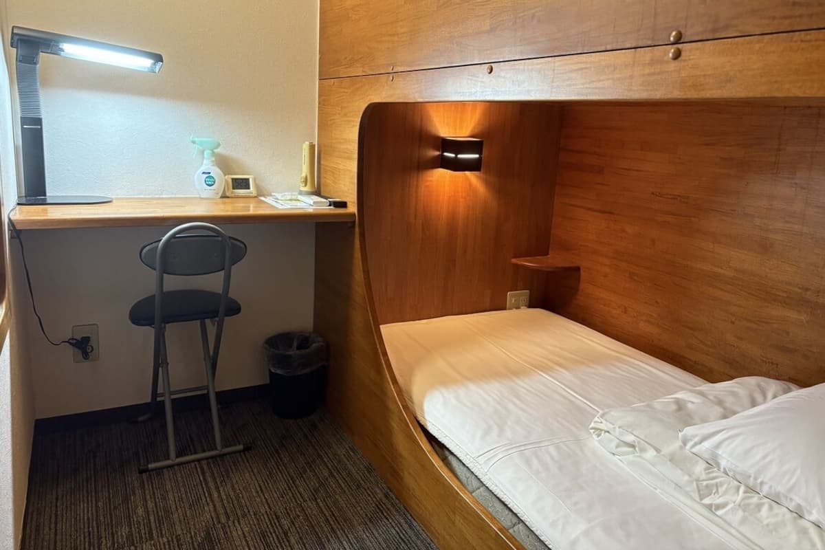 A compact room with a wooden headboard, a single bed, a desk with a chair, a desk lamp, and a small trash bin.