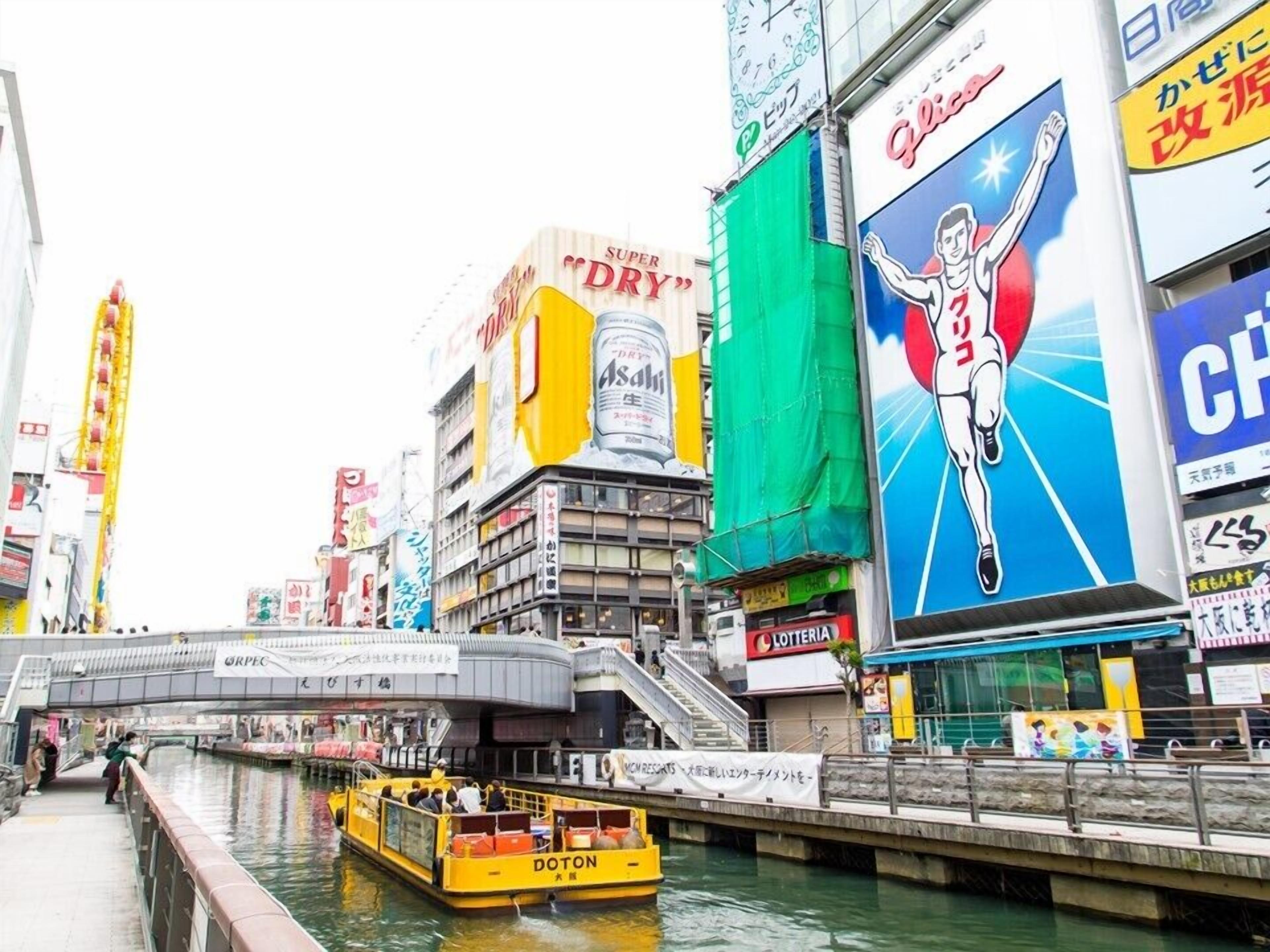 A busy urban street with a canal, a yellow boat, and various commercial buildings with signs and advertisements.