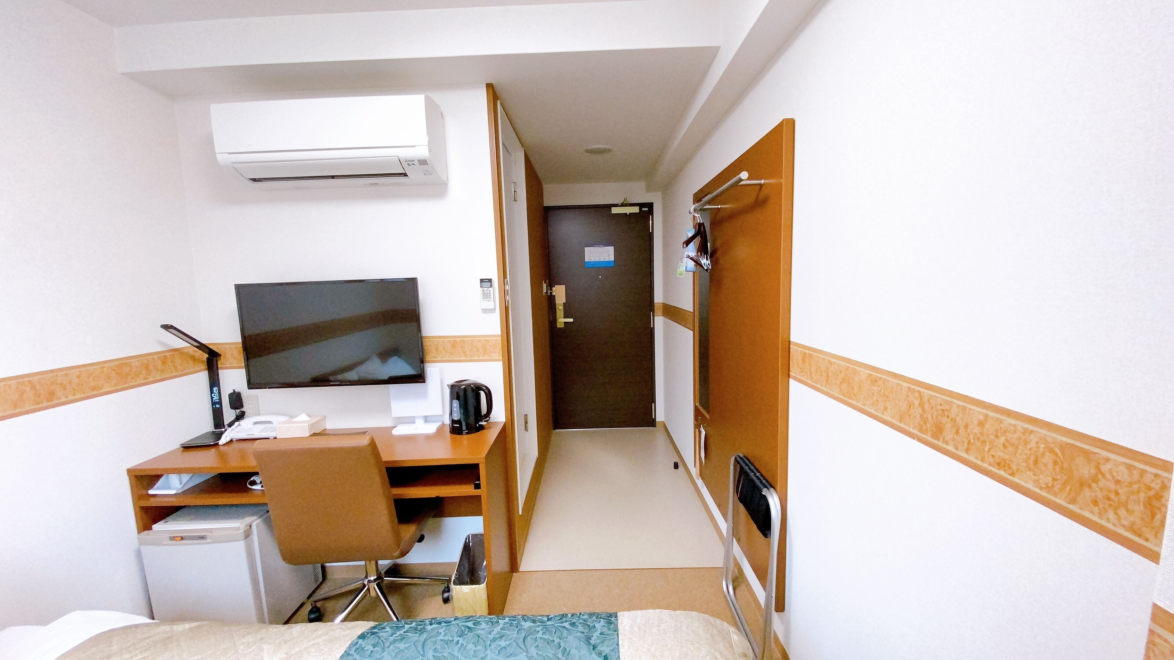 A compact hotel room with a bed, a desk with a computer, a TV, and a small kitchenette.