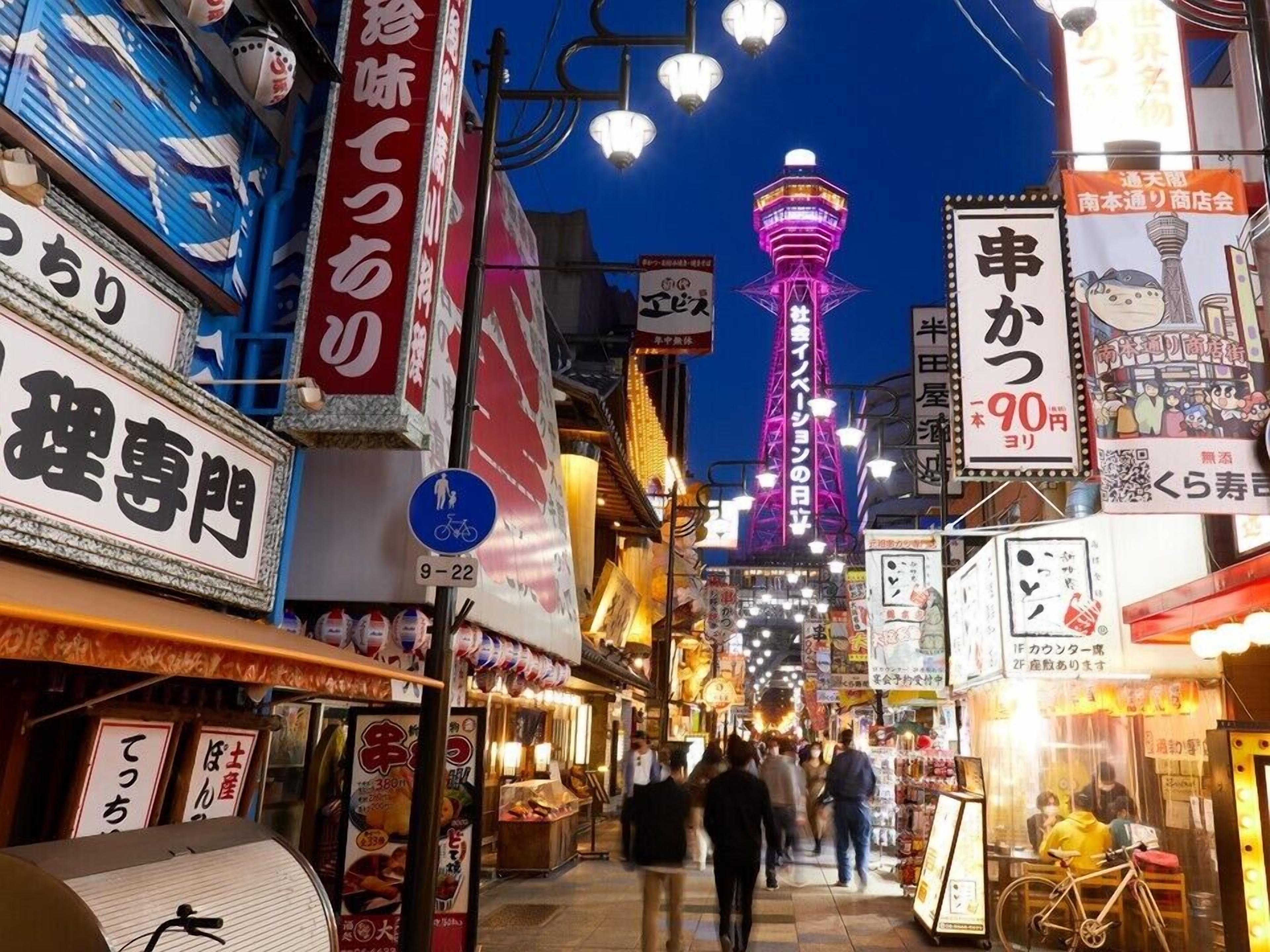 Una vivace scena di strada con insegne illuminate e una torre imponente sullo sfondo.