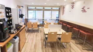 A cafeteria with wooden tables and chairs, a coffee machine, and a trash bin. - REX INN Namba (Osaka)