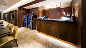 Zona de recepción de un hotel con mostrador de recepción, dos quioscos de autoservicio y un área de estar con mesas y sillas.