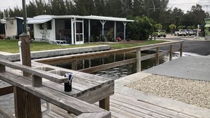 A wooden dock leading to a small house with a porch, a pond, and a gravel area.
