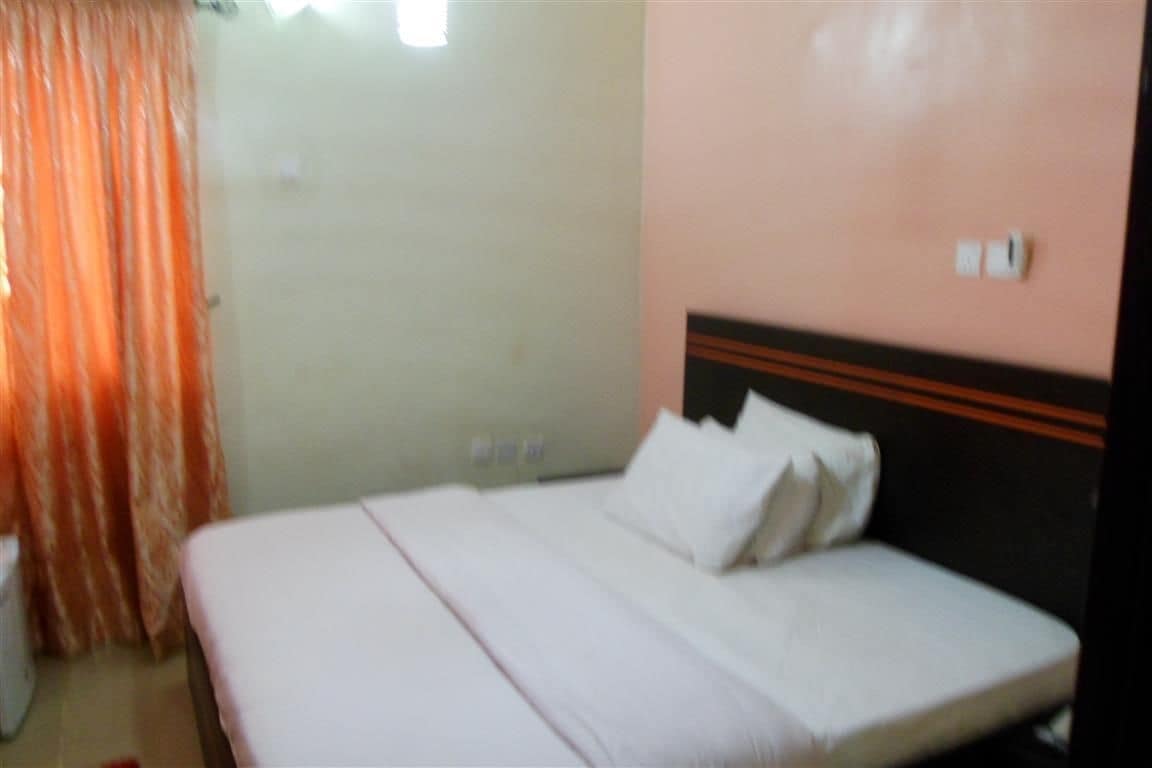 A bed with white bedding and a wooden headboard in a room with a plain wall and a window with curtains.