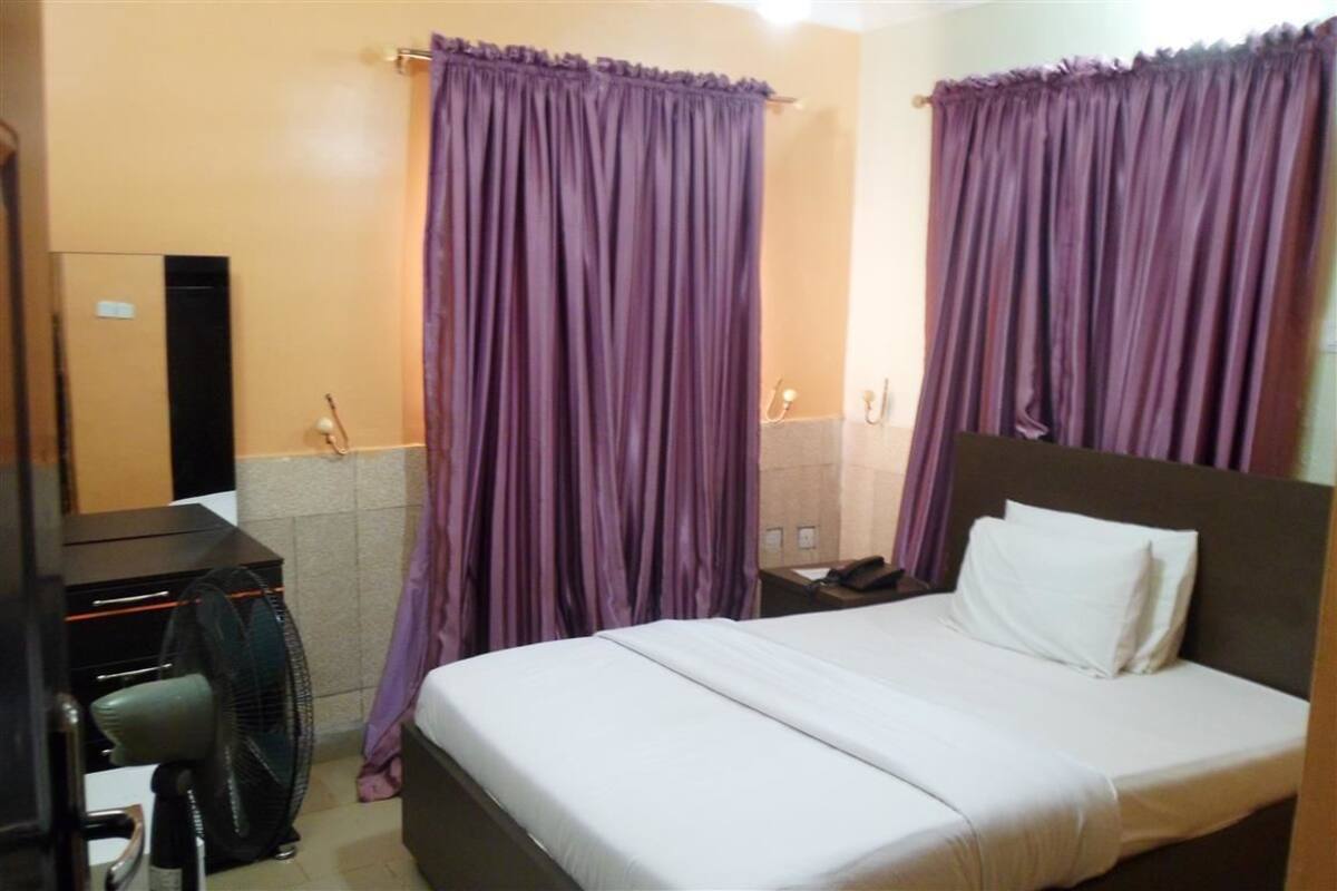 Quarto de hotel com cama, escrivaninha com telefone e cortinas roxas.