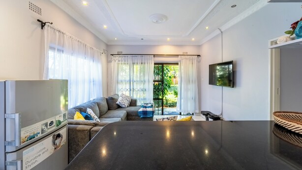 A modern living room with a large dark table, a sofa with cushions, a television, and a view of a garden through a glass door.