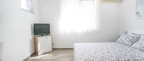 A bedroom with a bed, a TV on a wooden cabinet, a window with sheer curtains, and an air conditioner on the wall.