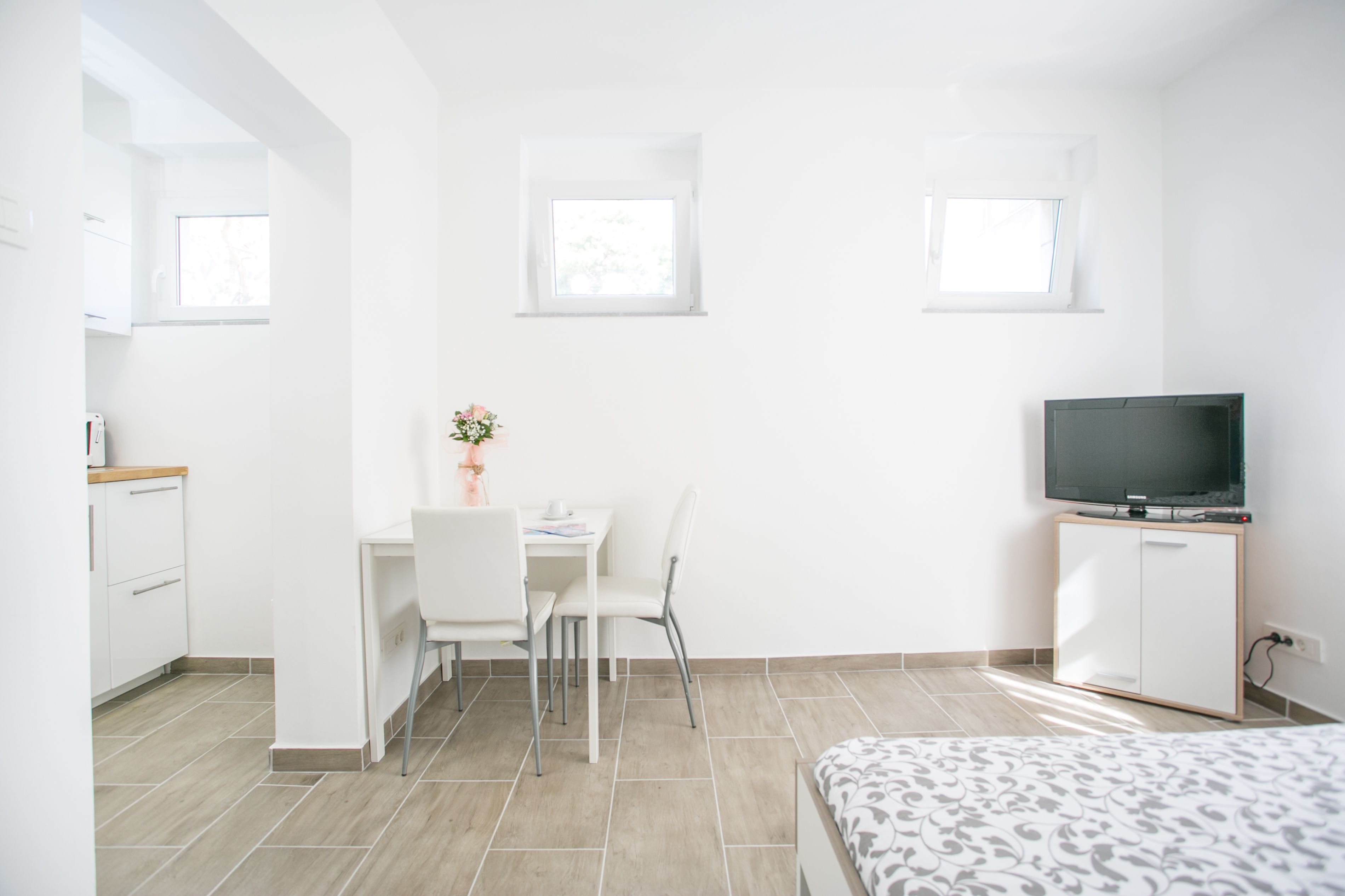 A modern, well-lit room with a bed, a TV on a stand, a desk with chairs, and a small kitchen area.