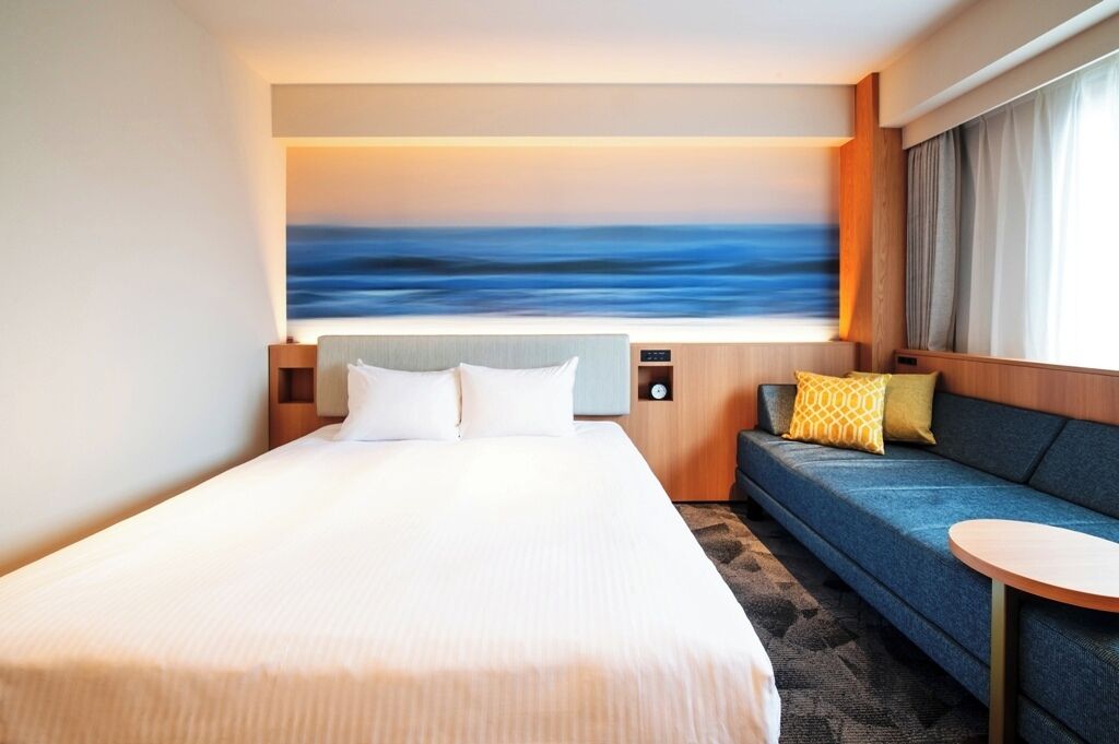 Una habitación de hotel con una cama grande, un sofá, una mesita y un cuadro de un paisaje marino en la pared.