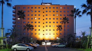 Un hotel con ingresso centrale, diversi piani e un parcheggio con diverse auto.