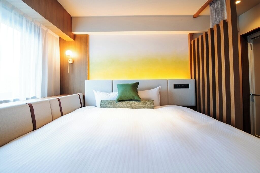 Ett hotellrum med en stor säng, en sänggavel med inbyggd luftkonditionering, ett nattduksbord och ett fönster med gardiner.