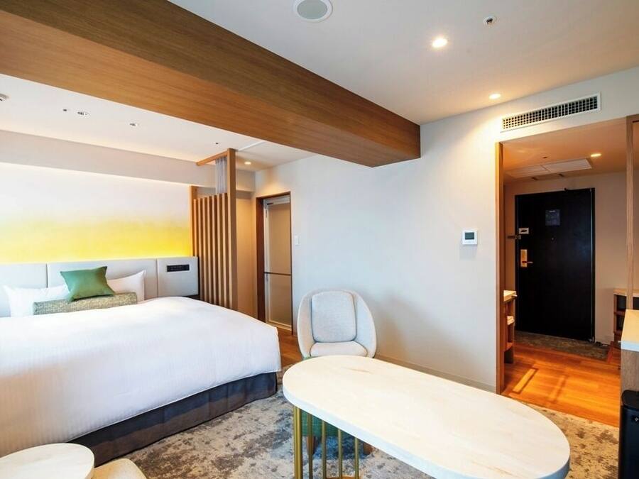 A modern hotel room with a large bed, a small table, and a chair. There is a wooden ceiling and a door leading to another room.
