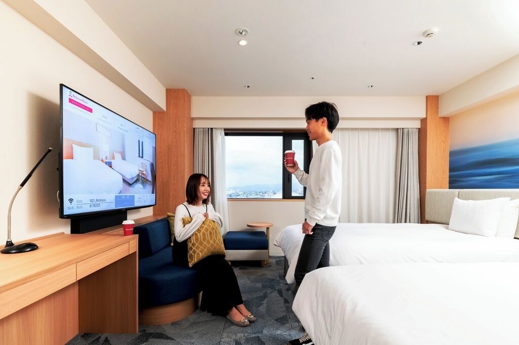 A hotel room with a bed, a TV on a desk, a sofa, and a window with a view.