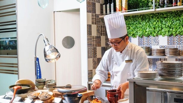 Uno chef in divisa bianca e con cappello da cuoco prepara cibo in una cucina con piastrelle a vista e diversi utensili e piatti sul bancone.