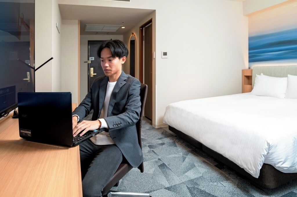 A hotel room with a bed, a desk with a laptop, and a view of the sea through the window.