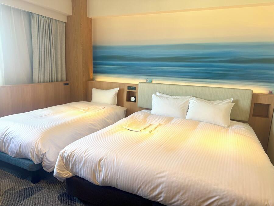 Habitación de hotel con dos camas, un cuadro de un mar tranquilo, un reloj y una pequeña estantería con un reloj.