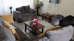 A living room with a grey sofa, a wooden coffee table with a flower vase, a grey armchair, and a small wooden table with a fan.