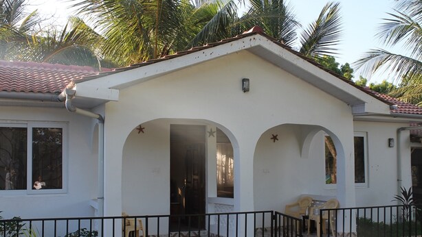 A two-story house with a tiled roof, a balcony with a railing, and a small garden area with palm trees.