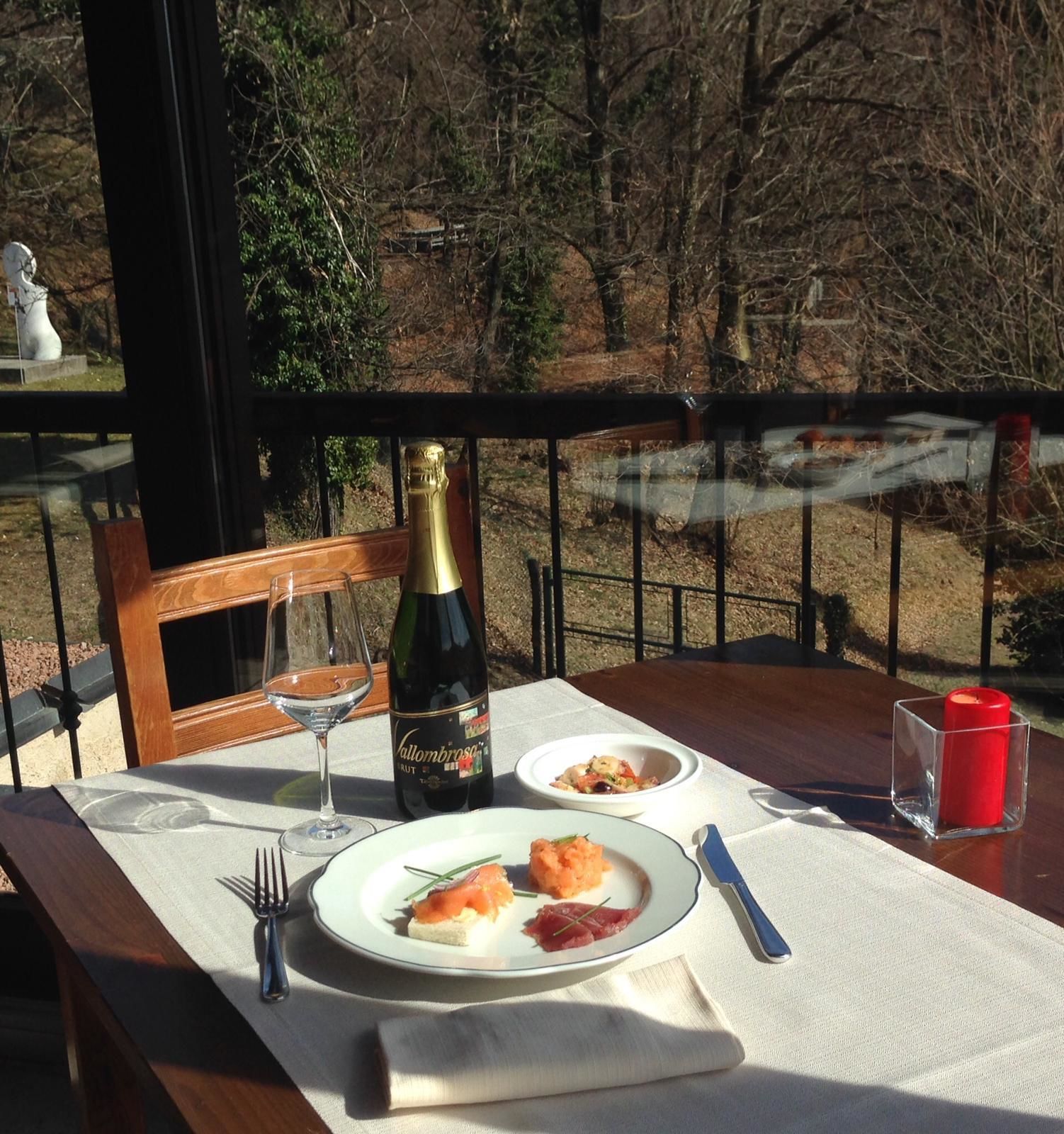 A table set with a bottle of wine, two glasses, a plate with food, a candle, and a fork on a balcony with a view of trees.