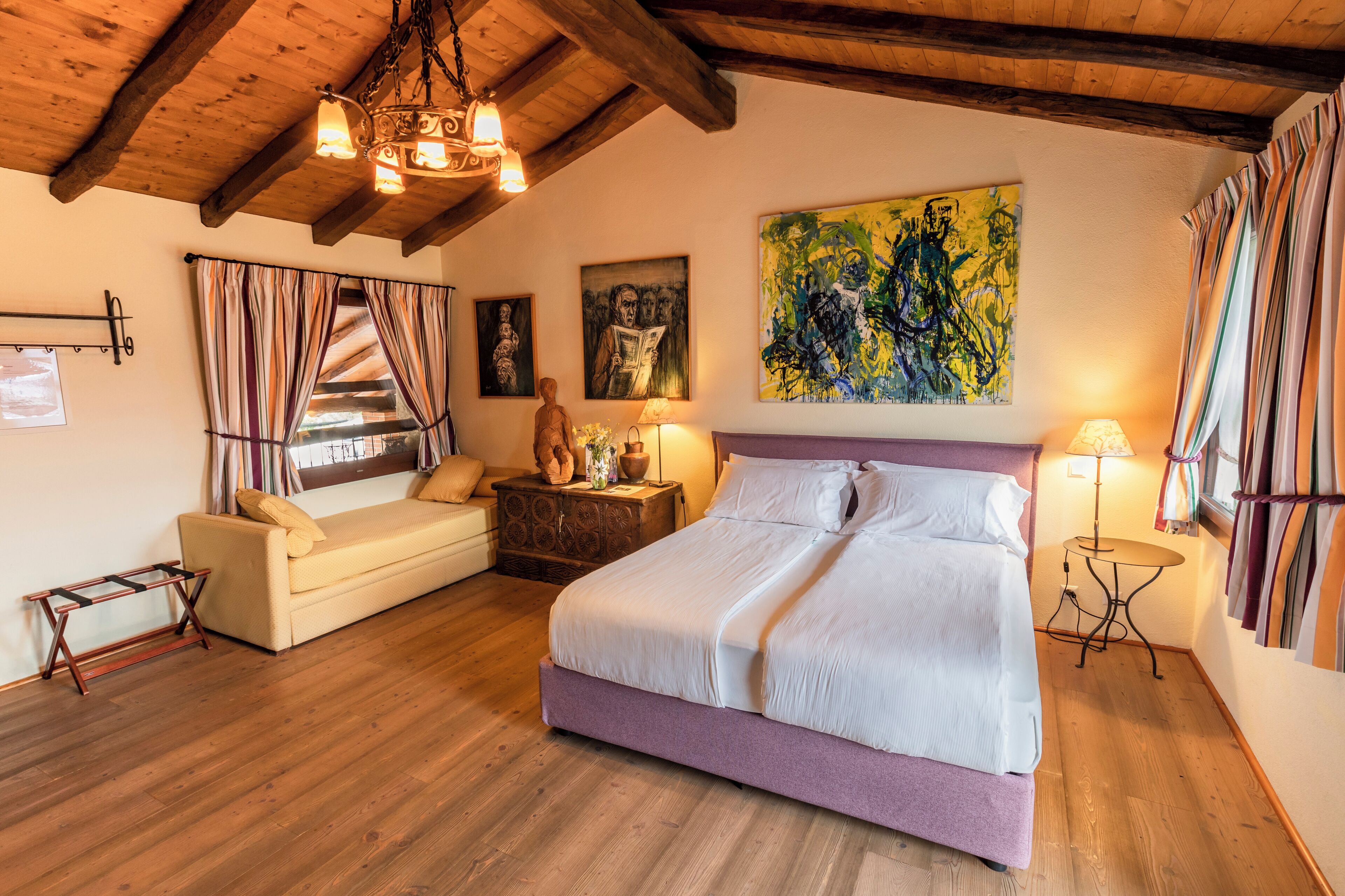 A bedroom with a bed, a sofa, a wooden floor, a chandelier, and artwork on the walls.