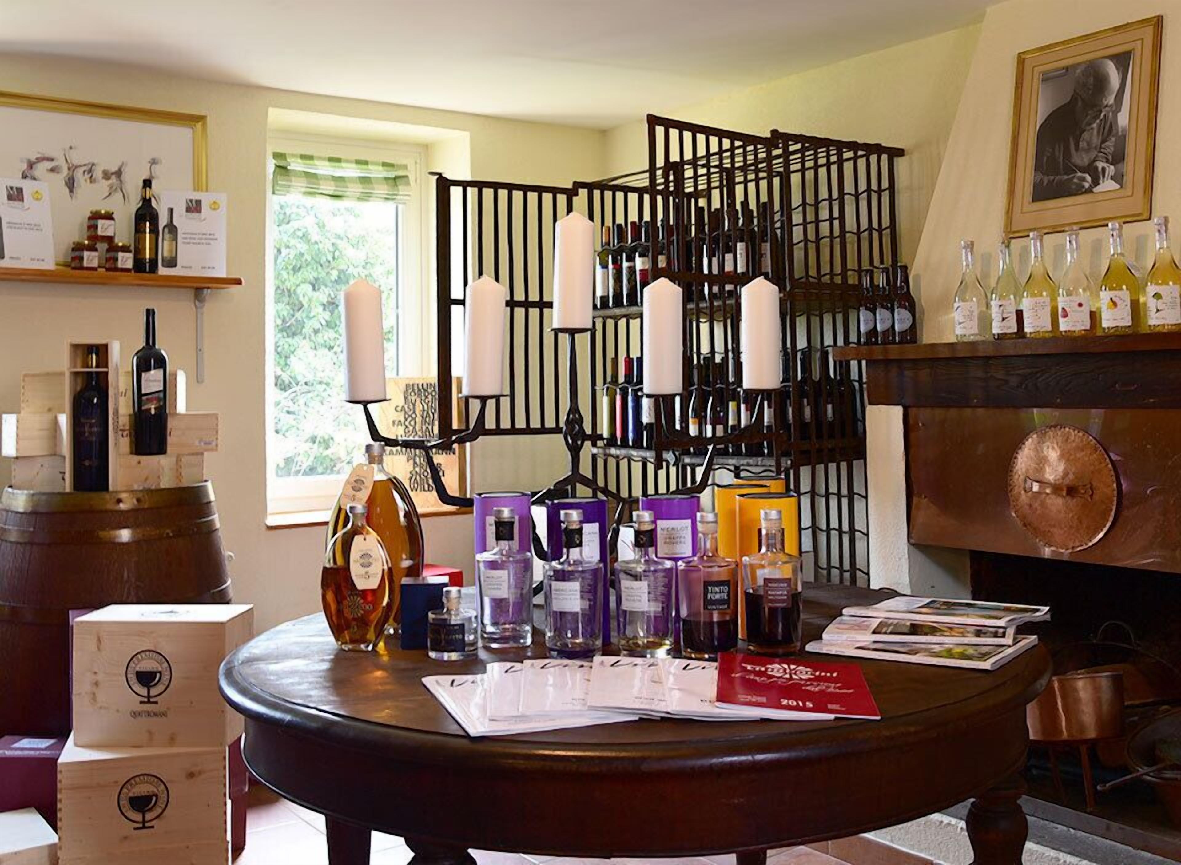 A room with a round table displaying various bottles and boxes, a black metal rack with candles, and a fireplace with wine bottles.