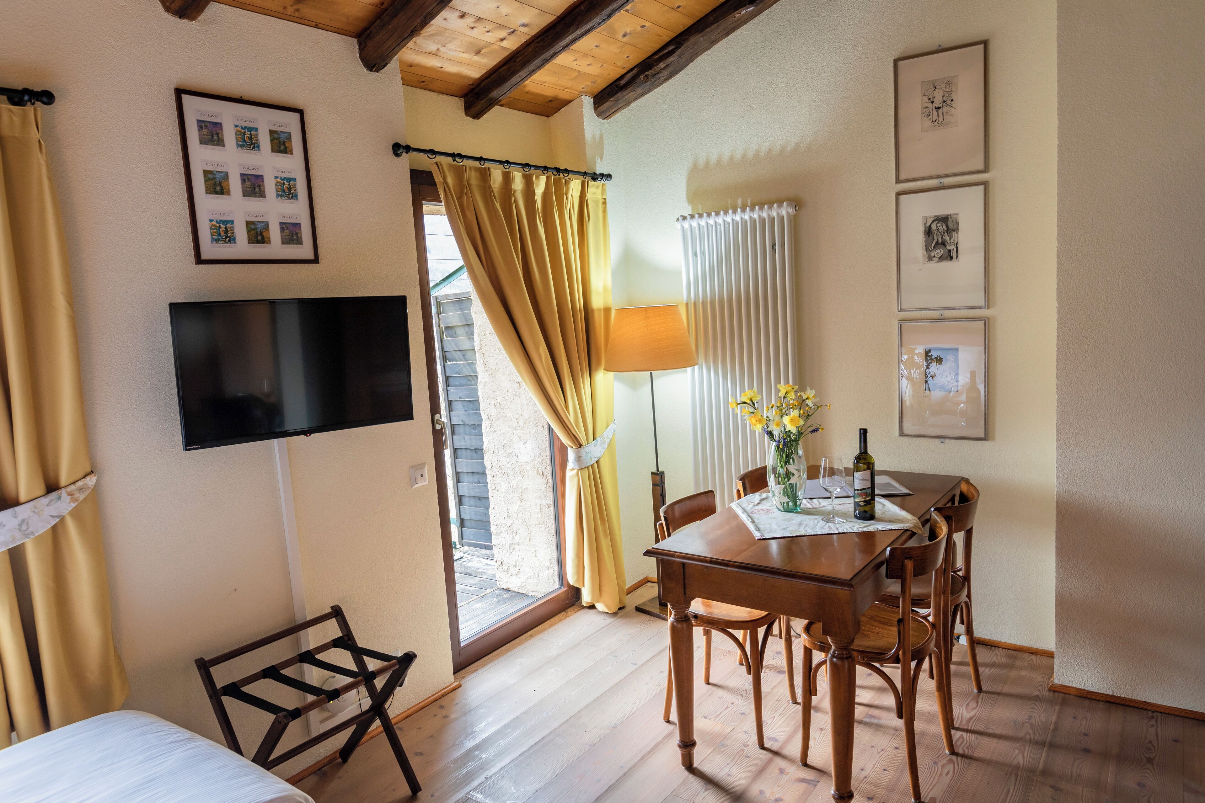 A room with a bed, a TV mounted on the wall, a dining table with chairs, and a window with curtains.