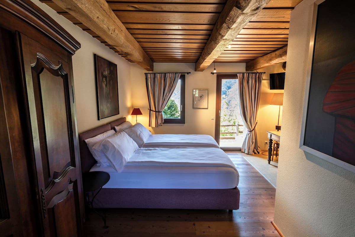 A bedroom with two beds, wooden ceiling beams, a window with curtains, and a painting on the wall.