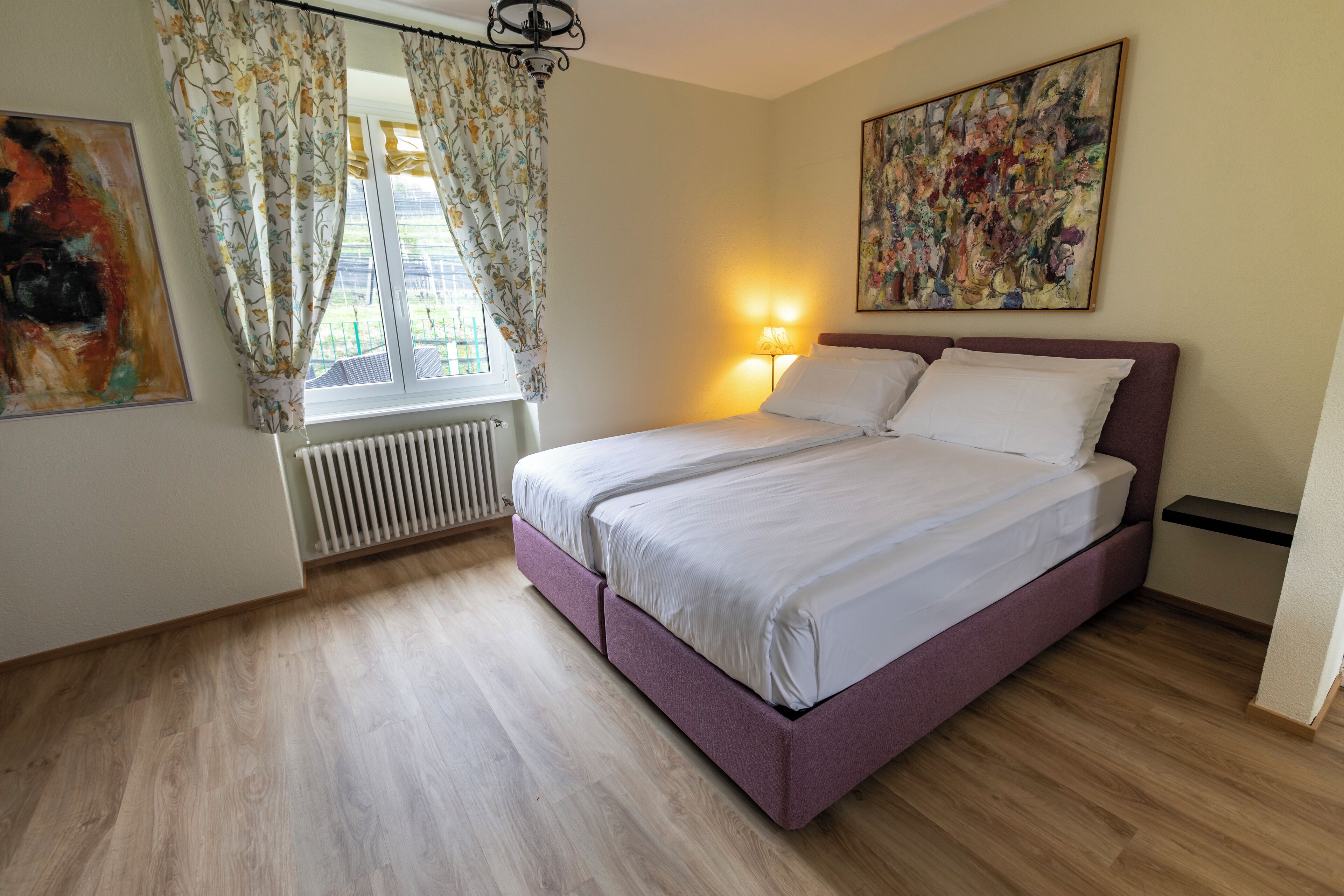 A bedroom with a bed, a window with curtains, a radiator, a wall-mounted lamp, and a painting.