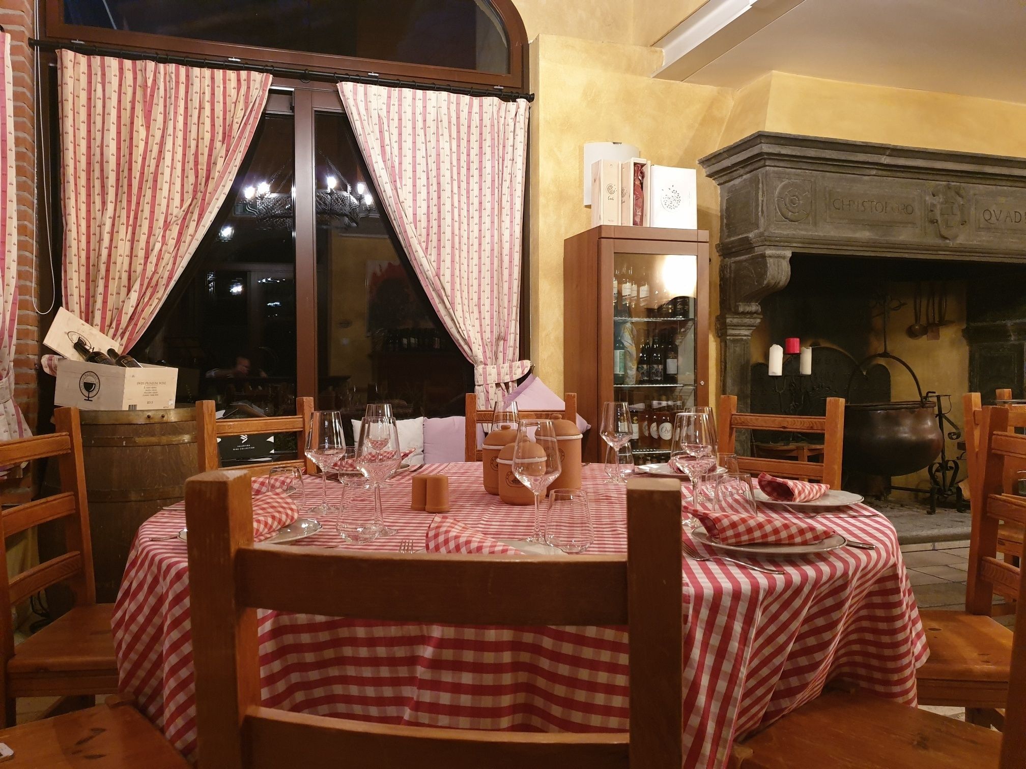 A dining area with a round table set for four, surrounded by wooden chairs. A large window with curtains is visible, and a fireplace with a pot is in the background.