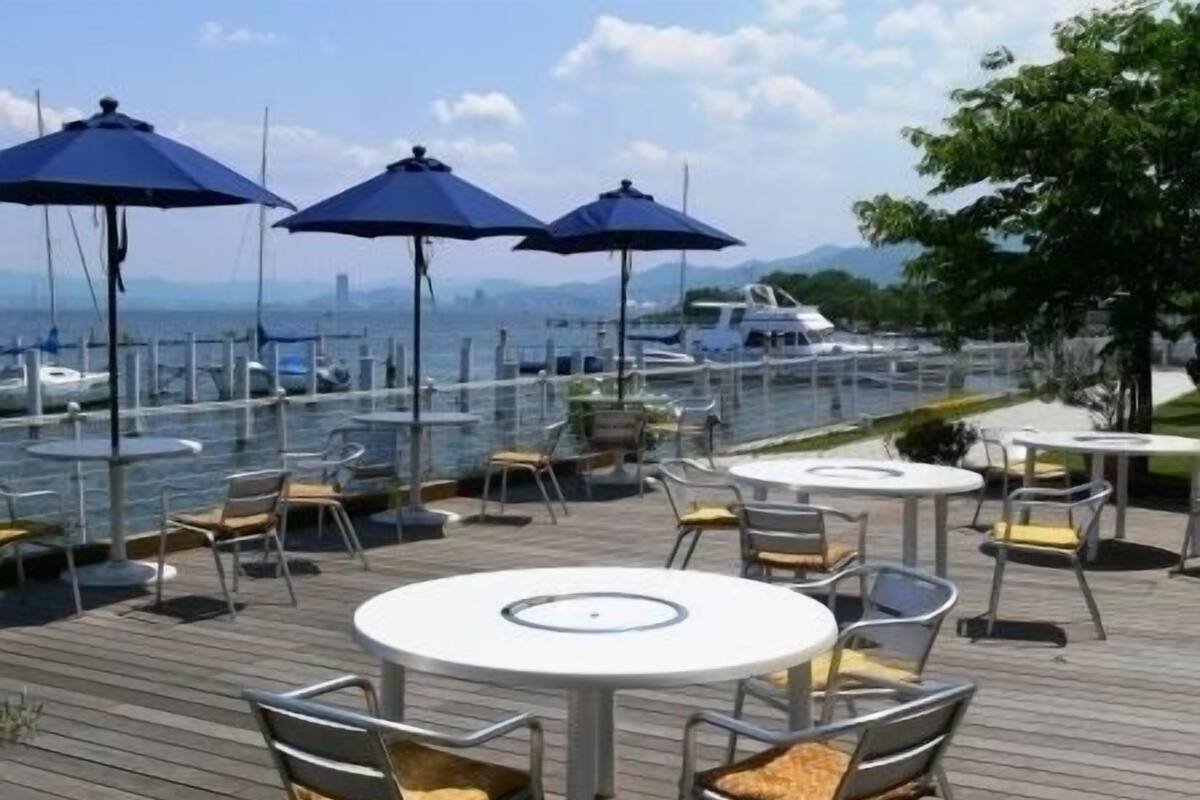 A waterfront patio with tables and chairs, umbrellas, and a view of boats and mountains.