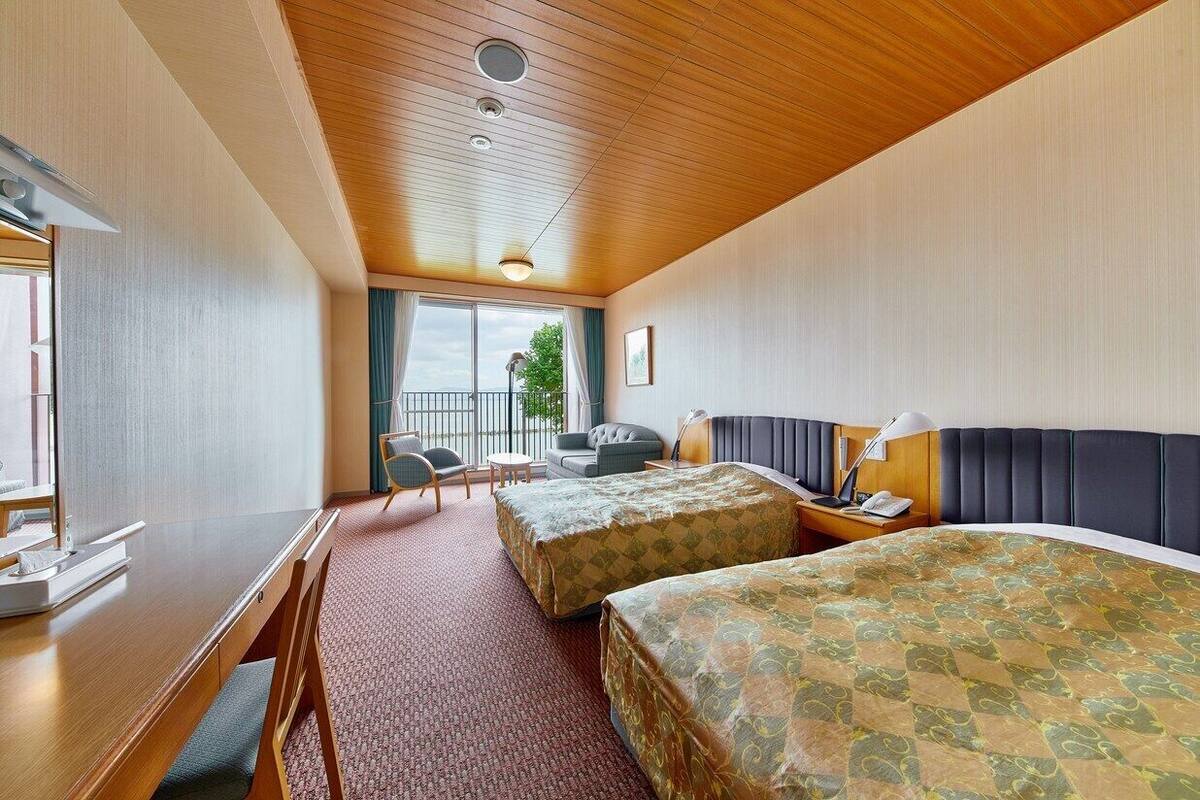 A hotel room with a wooden ceiling, two beds, a desk, a chair, a small table, and a balcony with a view.