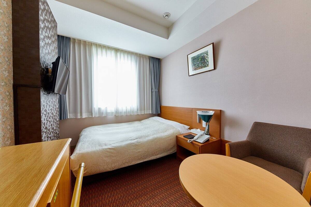 A hotel room with a bed, a desk, a chair, a nightstand with a lamp, and a window with curtains.