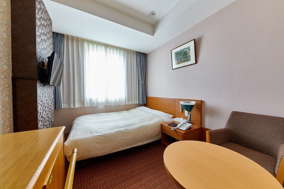 A hotel room with a bed, a desk, a chair, a nightstand with a lamp, and a window with curtains.