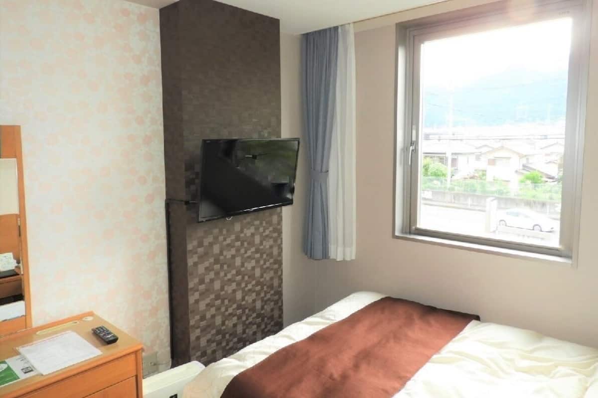 A hotel room with a bed, a TV mounted on the wall, a window with a view, and a desk with a remote control.