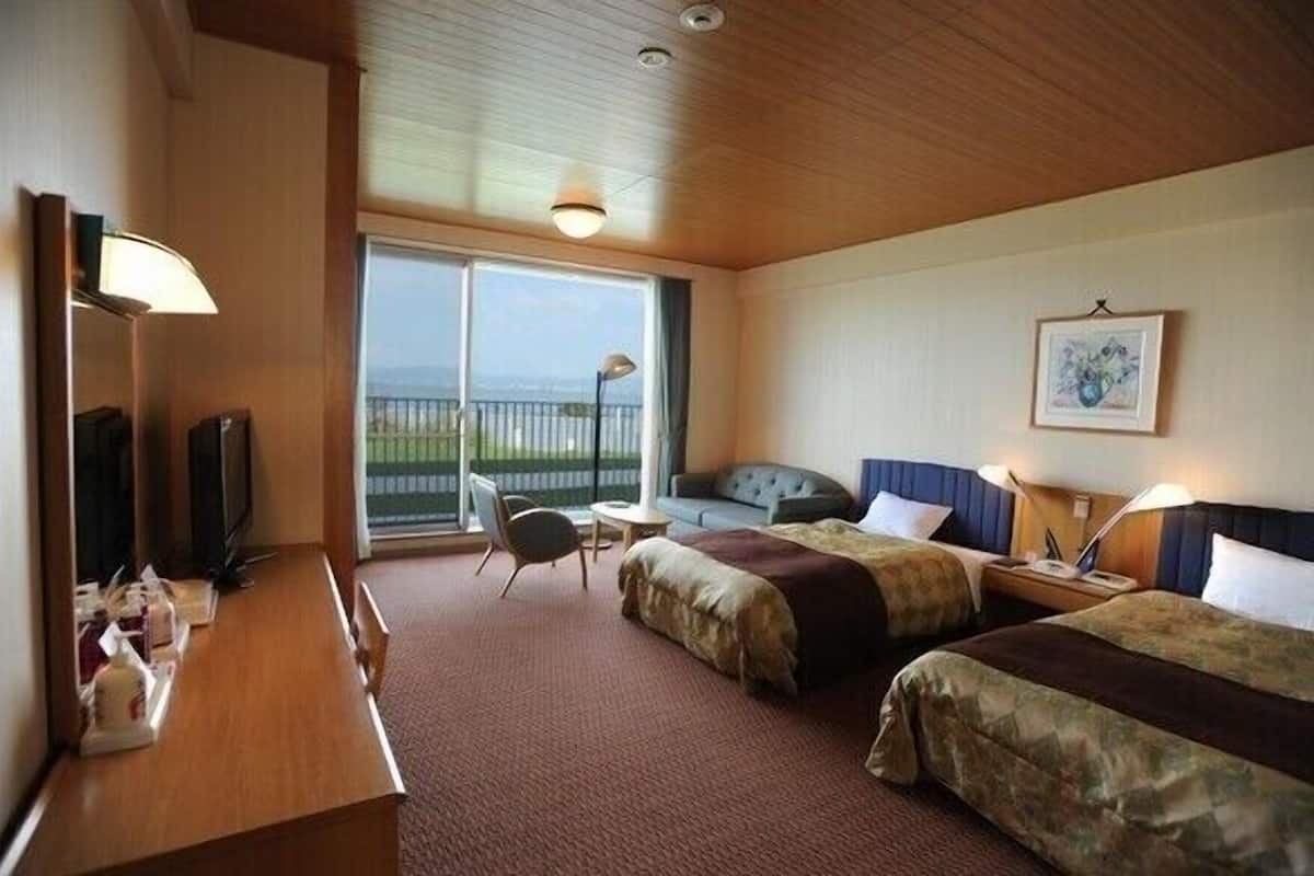 A hotel room with two beds, a desk, a chair, a TV, a balcony with a view, and a painting on the wall.