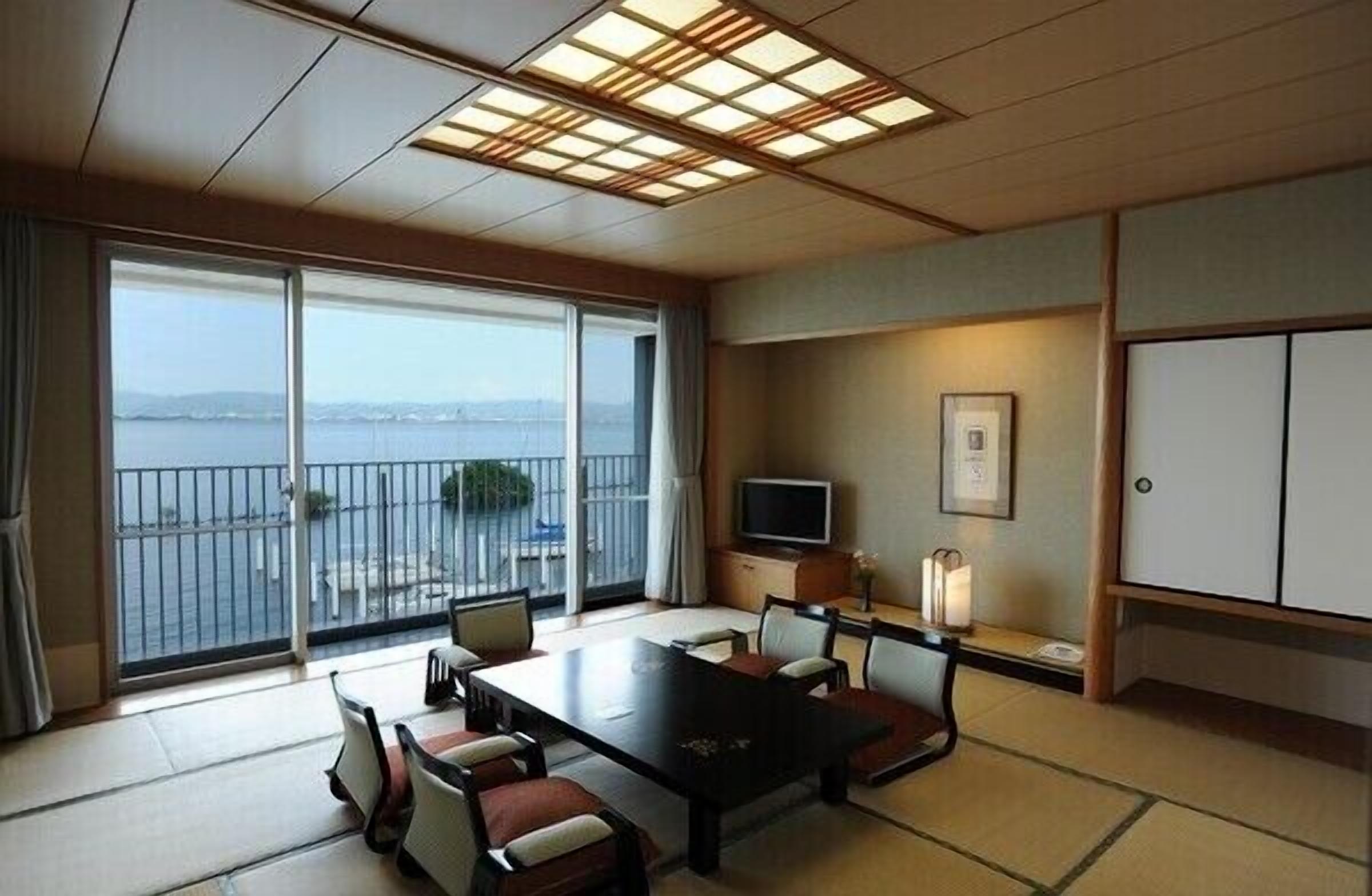 A hotel room with a large sliding glass door to a balcony, a flat-screen TV, a dining table with chairs, and a traditional Japanese-style cabinet.