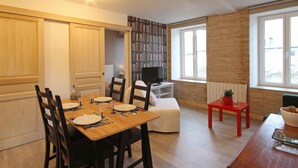 A dining area with a wooden table set for four, a bookshelf, a television, and a window with a view. - Literary Stay in Swiss Normandy with WIFI (Ségrie-Fontaine)