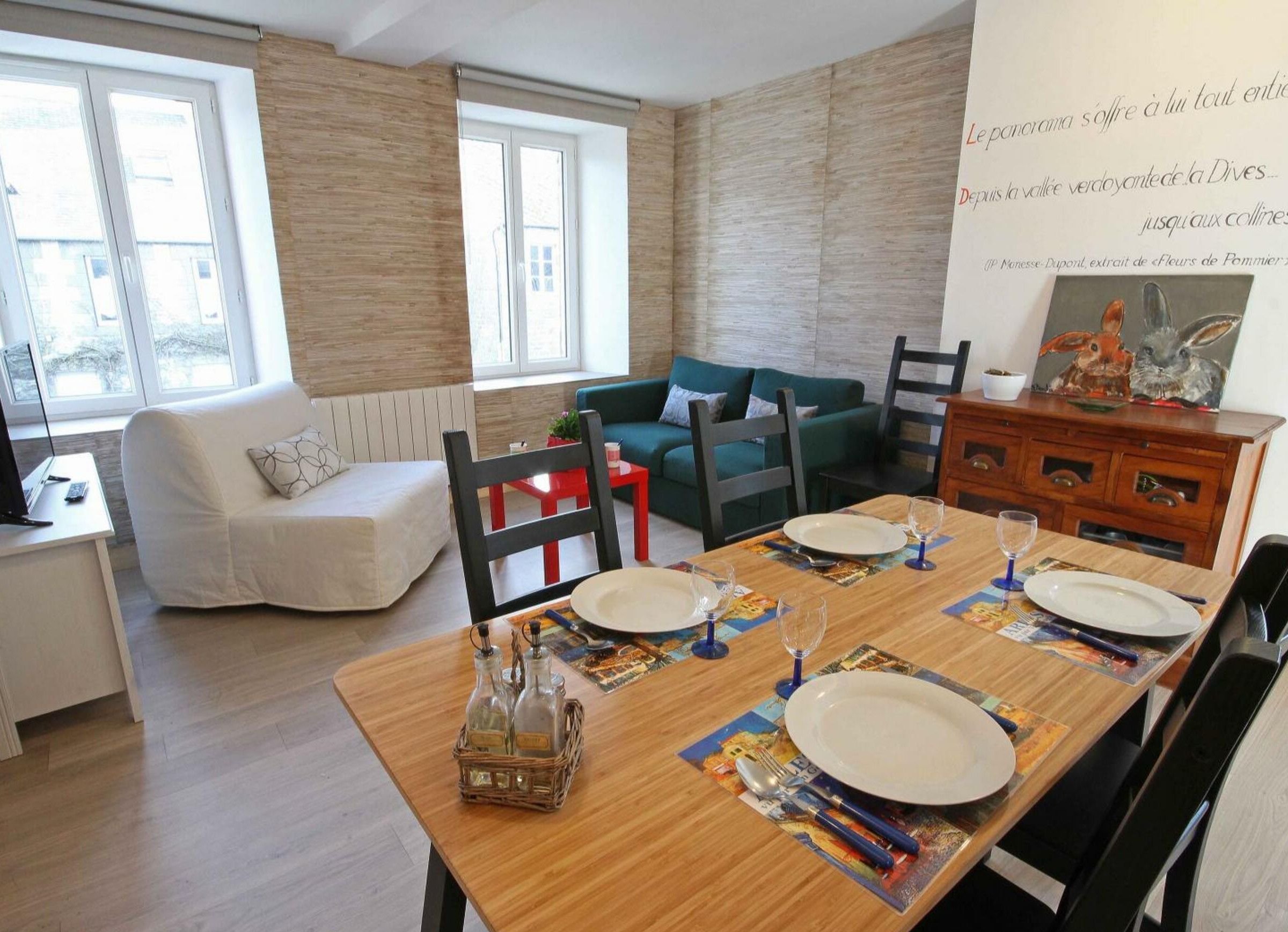 Un coin repas avec une table en bois dressée pour deux personnes, un canapé vert et un fauteuil blanc.