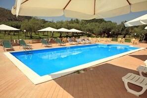 Ein Pool mit Liegestühlen und Sonnenschirmen, umgeben von einer Pflasterterrasse und Grünflächen.