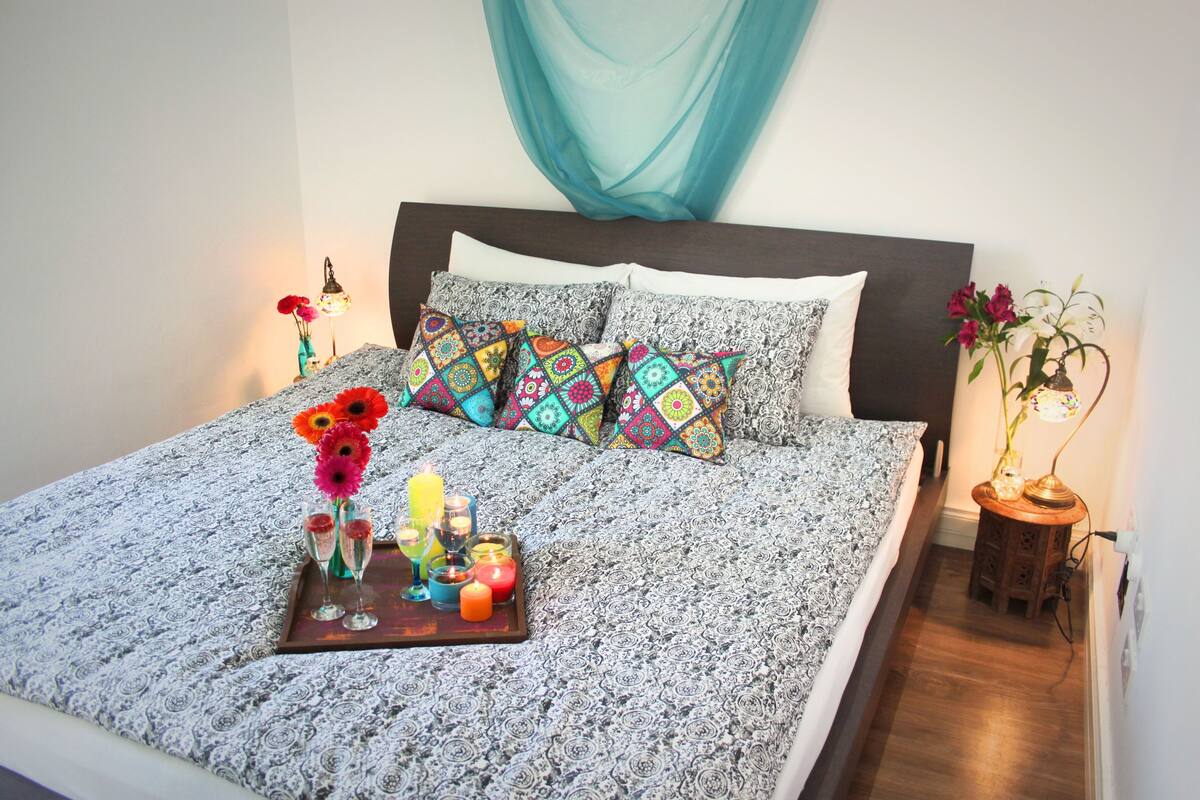 Una cama bien tendida con almohadones estampados, una bandeja con bebidas y flores, y una mesita con lámpara y flores.