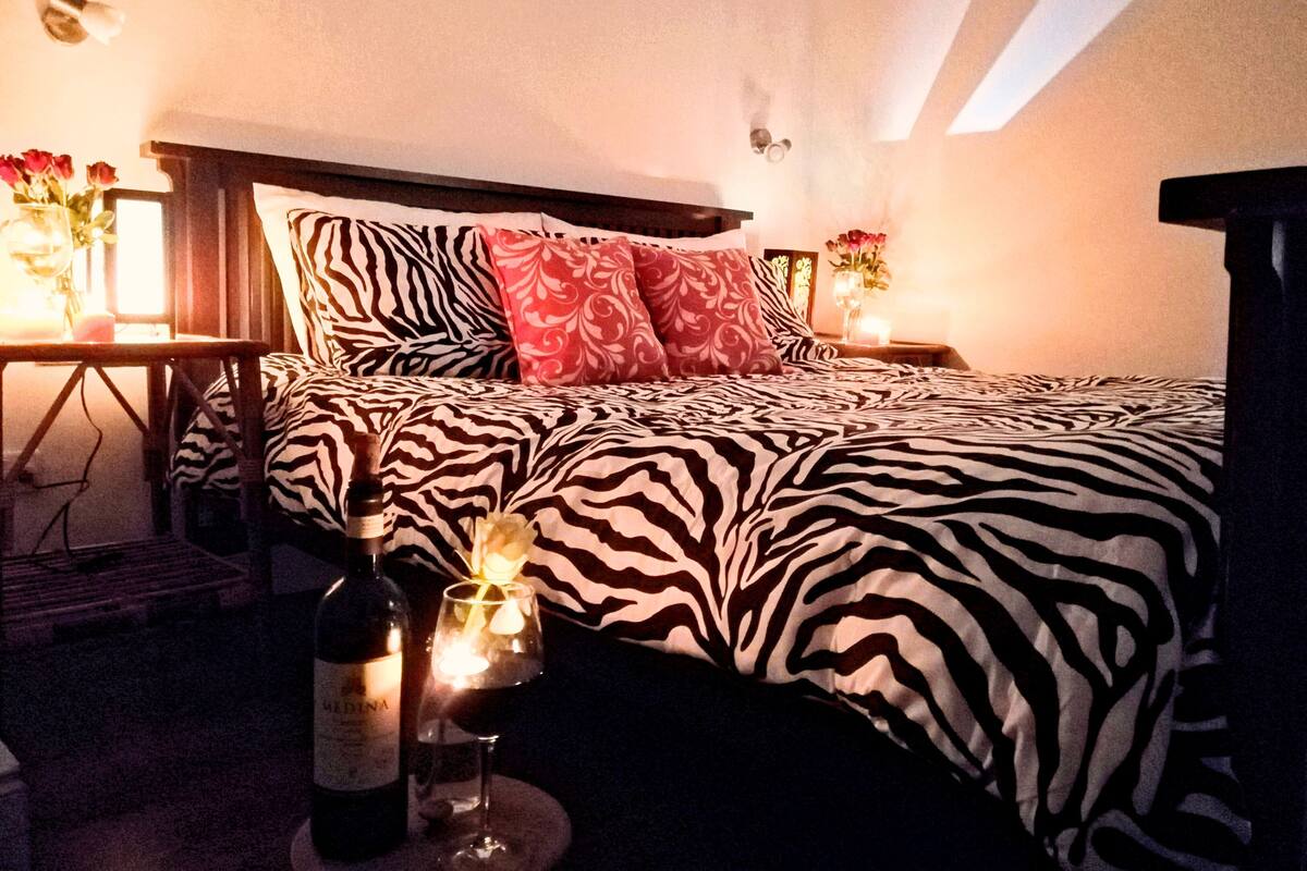 Un dormitorio con una colcha de estampado de cebra, una mesita de noche con un jarrón de flores y una botella de vino sobre una pequeña mesa.