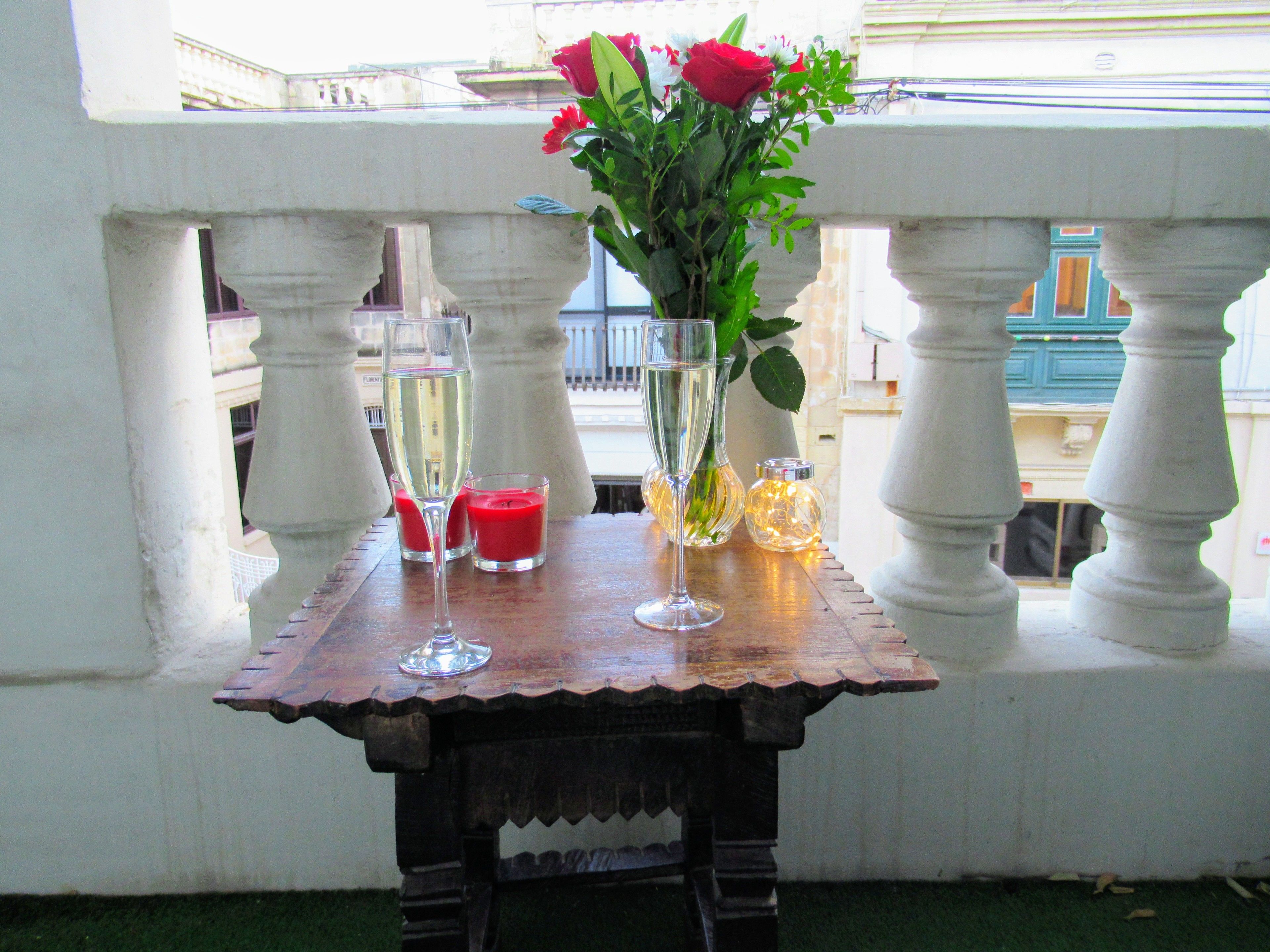 A table with two champagne flutes, a vase with red flowers, and two lit candles on a balcony railing.