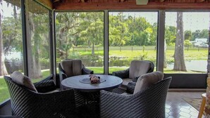 Un patio cubierto con mobiliario de mimbre y una mesa redonda, con vistas a un paisaje natural con árboles y una masa de agua.