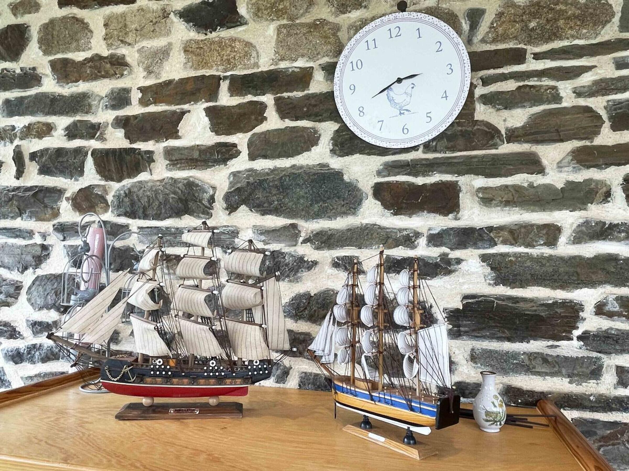 Un modelo de barco sobre una mesa de madera, frente a una pared de piedra, con un reloj encima.