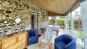 A room with a stone wall, wooden ceiling, and a table with chairs. There is a clock on the wall and a window with a view of greenery.